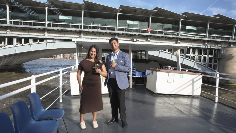 Two guests pose with drinks on the open deck as the boat passes under Blackfriars Bridge